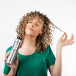 Cargar imagen en el visor de la galería, Chica con pelo rizado y rubio sujetandose un mechón de pelo con una mano y con la otra un bote de COOL BLONDE SHAMPOO