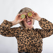 Cargar imagen en el visor de la galería, Chica rubia platino con pelo corto sonriendo con boca abierta sujetando un kiwi partido en cada mano delante de los ojos