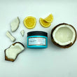Cargar imagen en el visor de la galería, Mascarilla hidratante y reparadora A Must Coco Mask tumbada en el suelo de frente rodeada de limones, piezas de coco y medio coco hacia arriba.