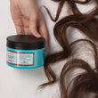 Cargar imagen en el visor de la galería, Mano sujetando mascarilla reparadora THE GLAM MASK con mechones de pelo castaños ondulados alrededor