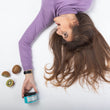 Cargar imagen en el visor de la galería, Chica castaña con pelo largo boca abajo tumbada en el suelo mirando y sujetando con la mano mascarilla nutritiva THE GLAM MASK con tres kiwis alrededor