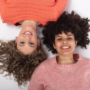 Dos chicas tumbadas una en cada dirección mirando a cámara y sonriendo. Una con pelo rizado rubia y otra con pelo moreno rizado afro