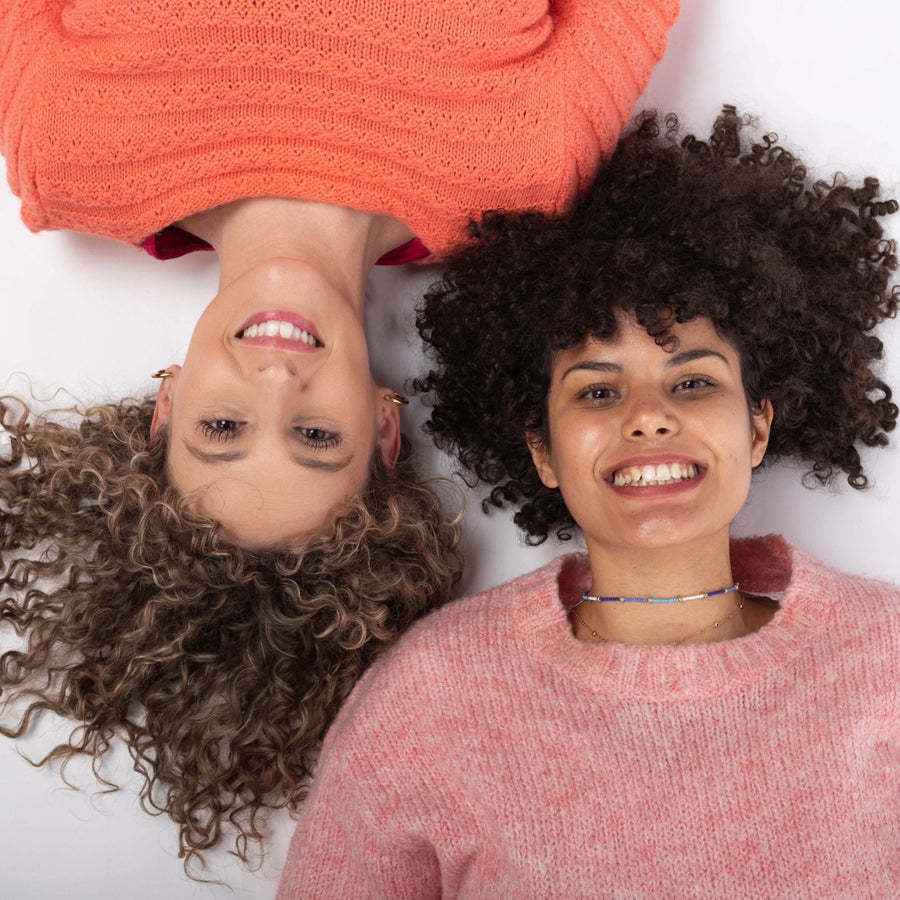 Dos chicas tumbadas una en cada dirección mirando a cámara y sonriendo. Una con pelo rizado rubia y otra con pelo moreno rizado afro