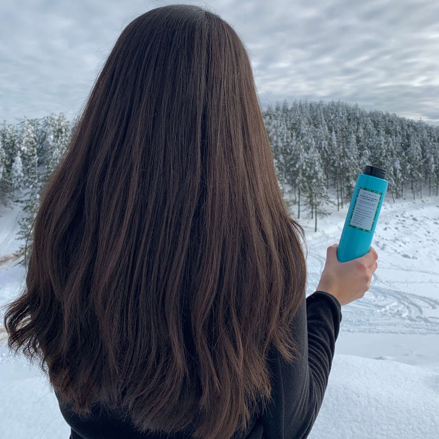 Chica morena con pelo largo, abundante y cuidado de espaldas mirando a la nieve sujetando champu natural sin sulfatos Marine Plasma Shampoo