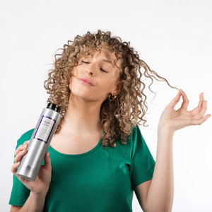 Chica con pelo rizado y rubio sujetandose un mechón de pelo con una mano y con la otra un bote de COOL BLONDE SHAMPOO 