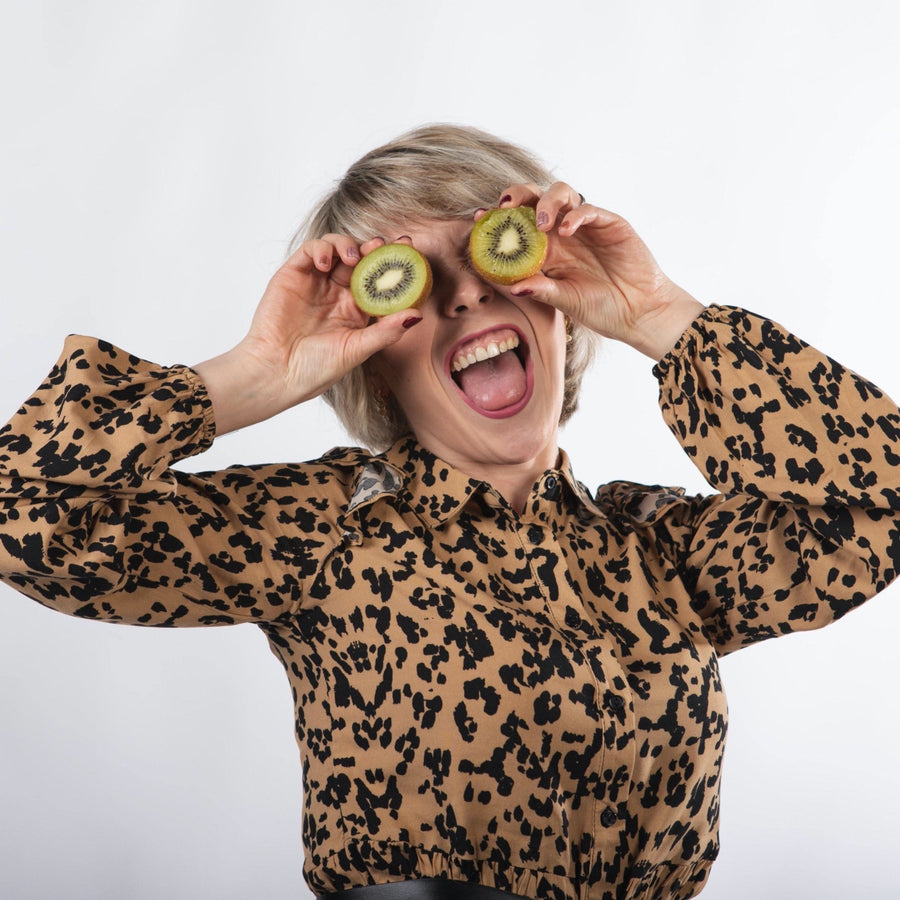 Chica rubia platino con pelo corto sonriendo con boca abierta sujetando un kiwi partido en cada mano delante de los ojos