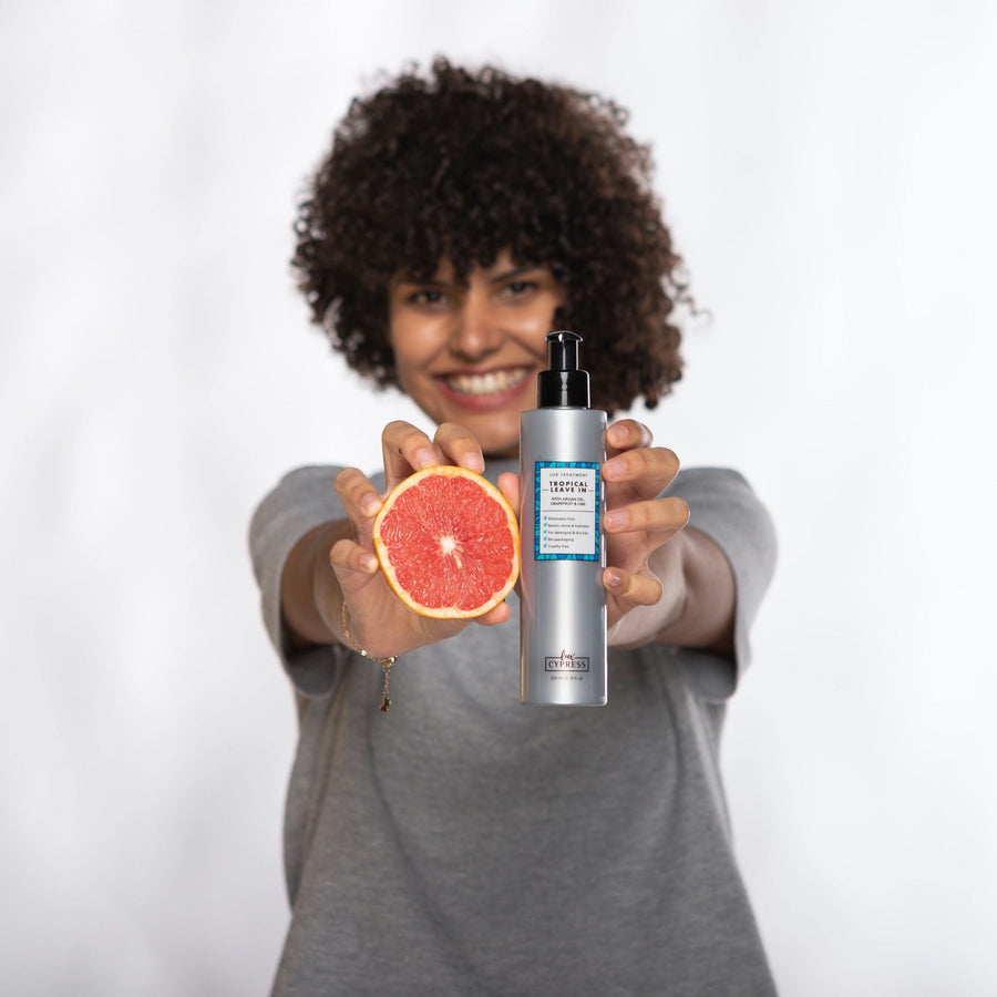 Chica con pelo rizado afro sonriendo sujetando un pomelo y tratamiento sin aclarado TROPICAL LEAVE IN con cada mano